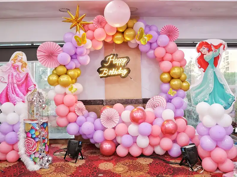 A pink and gold birthday backdrop with paper fans, princess cut-outs and a Happy Birthday sign.