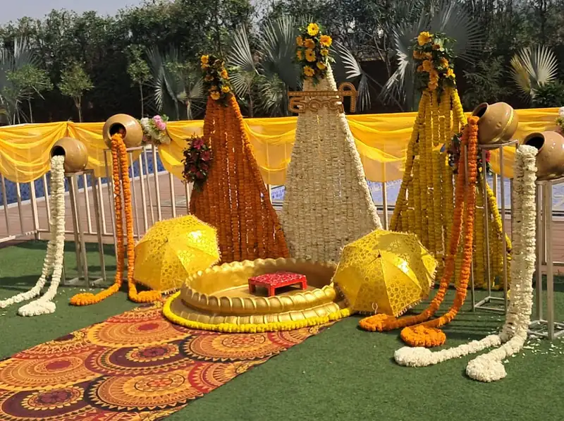 A haldi setup on a lawn with marigold strings, gold pots and a low decorated seat.