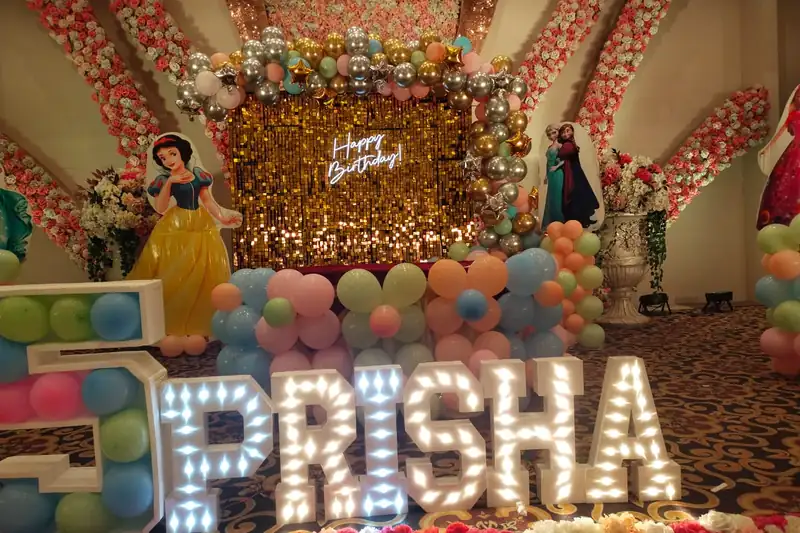 Bulb letters spelling a child's name in front of a gold shimmer panel with pastel balloons.